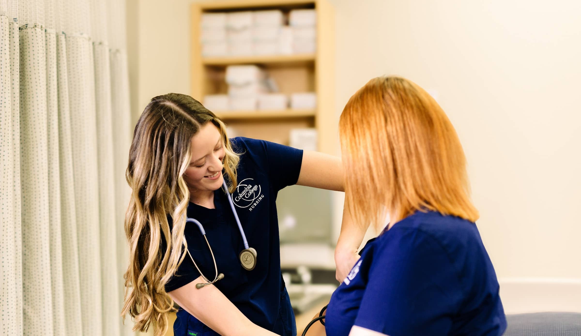 Female nursing student, wearing CC blue scrubs and with a stethoscope around her neck, takes the blood pressure of a seated instructor.