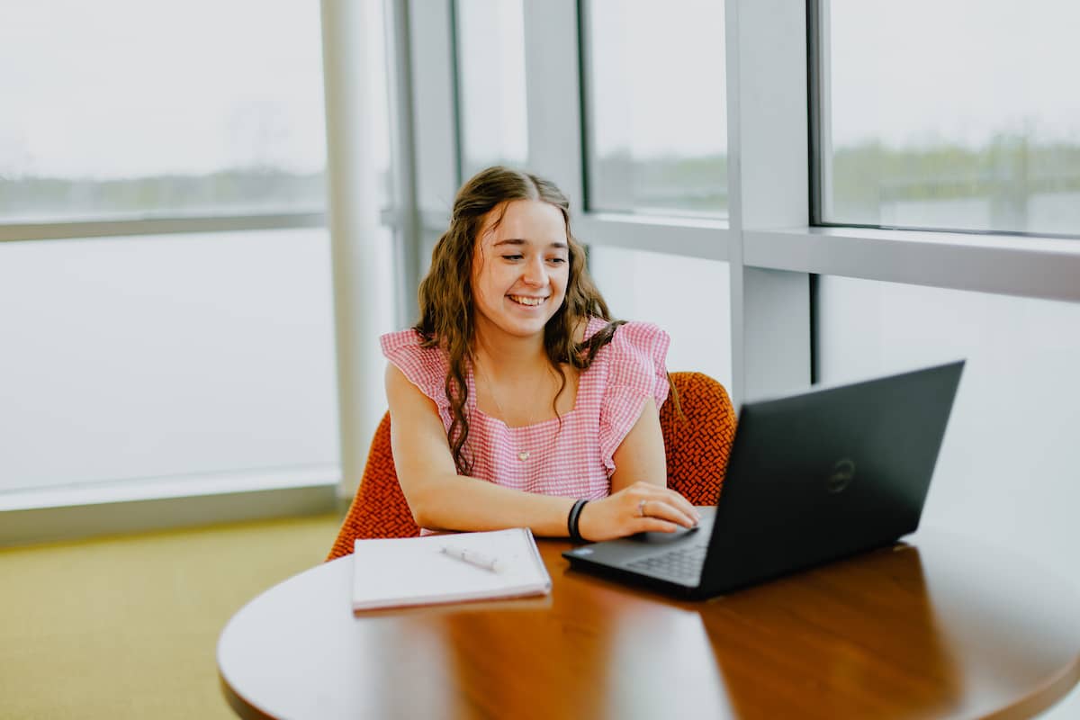 Student sitting at table, smiling while reviewing work on a laptop.