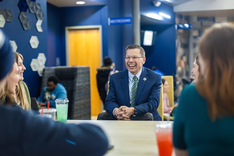 Dr. Toby Arquette, wearing a blue suit and stiped green and gray tie, sitting in Atkins-Holman commons, sharing a laugh with four students.