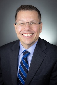 Headshot of Dr. Toby Arquette, wearing a dark suit, dark/light blue tie.