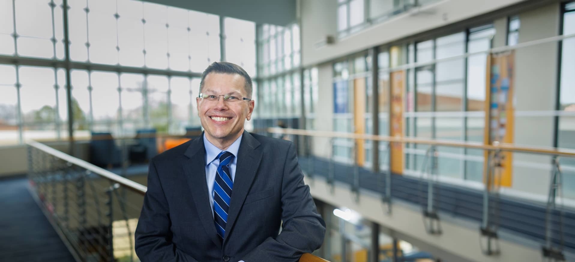 Dr. Toby Arquette, CC's 19th president, standing on the upper level of Atkins-Holman Student Commons, wearing a dark suit, a light blue shirt, and a dark/light blue striped tie.