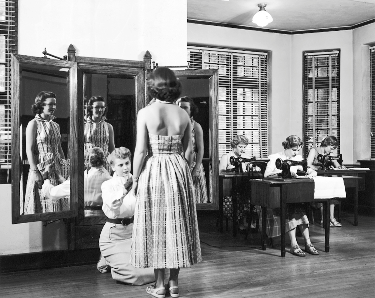 Historical image of a student kneeling, hemming the dress of a student who is standing in front of mirrors in a sewing class.
