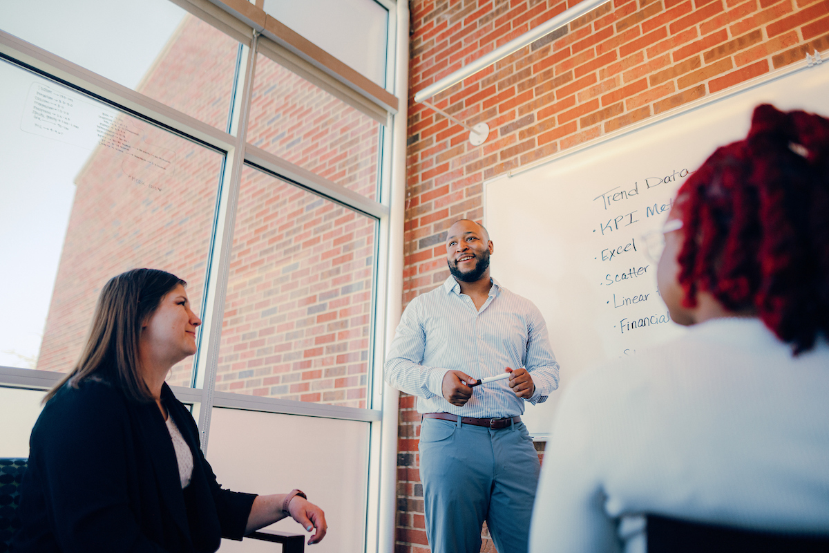 Two people sitting, and one person standing in front of a whiteboard, involved in conversation.