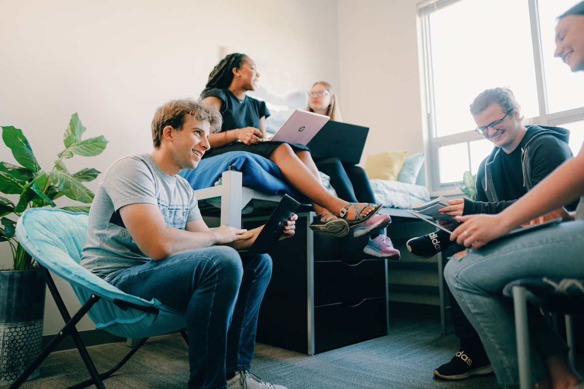 Five students sitting around having a discussion, with to students engaged in a conversation and using laptops.