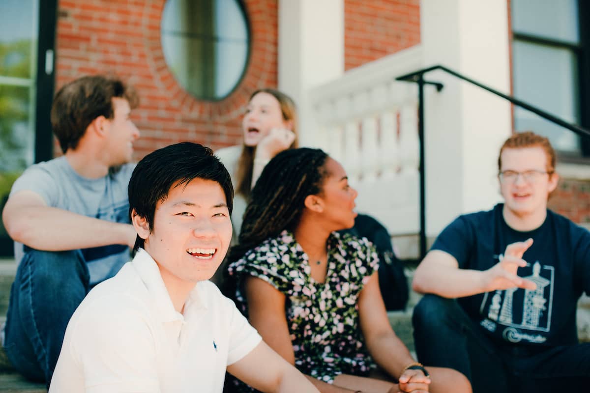 Students sitting on cement steps outside on main campus, some in discussion, one turning to smile at the camera.