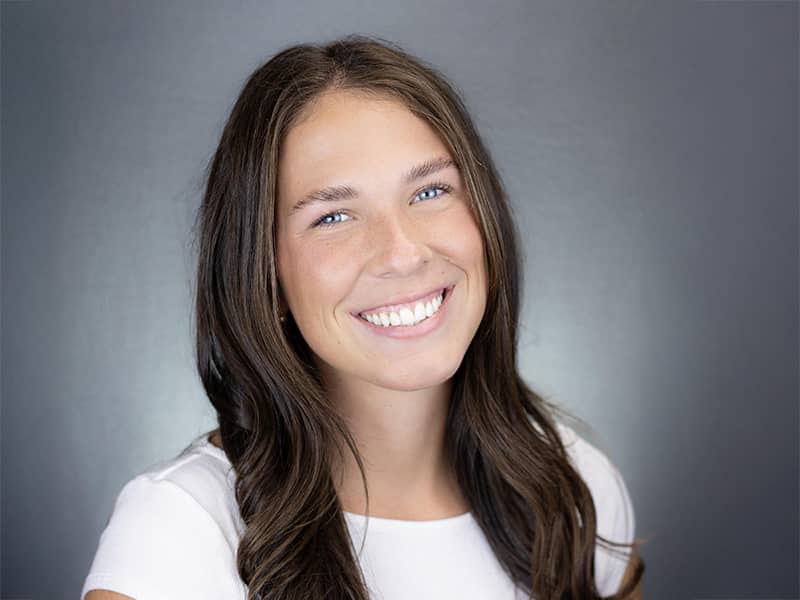 Headshot of Caleigh Huot, long brown hair hanging down over a white t-shirt, smiling at the camera.