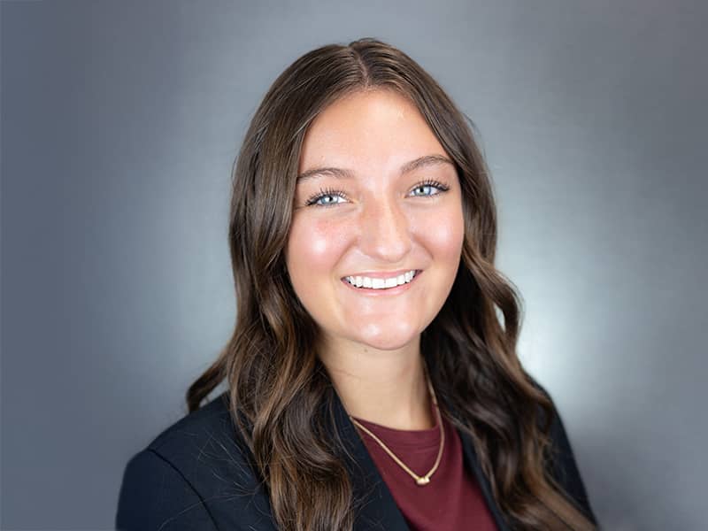 Headshot of Emiley Dial, long brown hair hanging down over a maroon shirt and dark jacket, smiling at the camera.