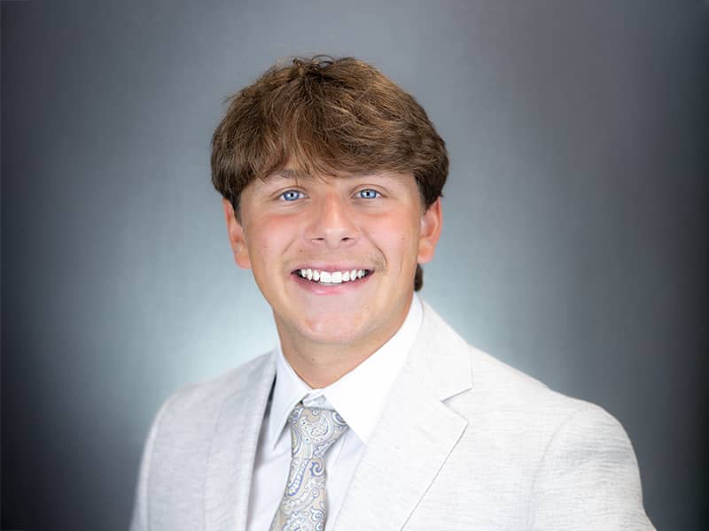 Headshot of Hunter Shoulta, wearing a light suit jacket, patterned tie, and white dress shirt, smiling at the camera.