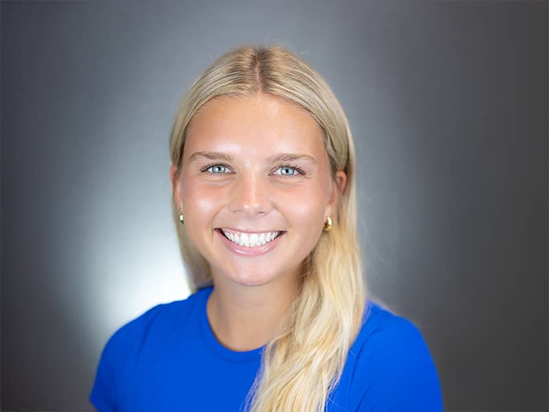 Headshot of Izzy Cole, long blond hair hanging down over a bright blue t-shirt, smiling at the camera.