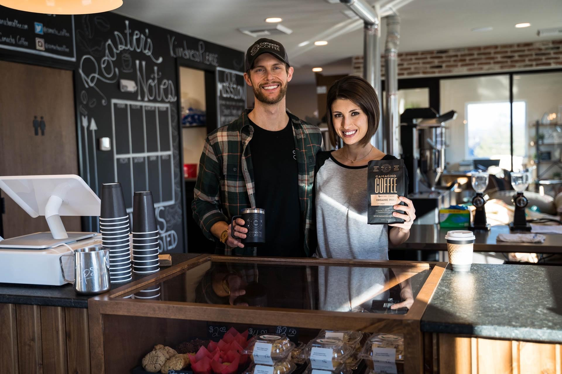 Jesse Walter wearing a plaid shirt and baseball cap, standing next to his wife, Megan, in her t-shirt, smiling at the camera while holding bags of their coffee.